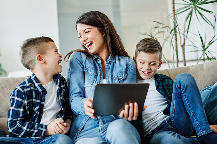Client Center - Happy Mother is Laughing With Her Two Sons While Sitting on a Sofa Using a Tablet at Home